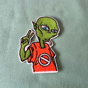 Könnte beinhalten: Grüner Außerirdischer mit blauen Augen, der ein orangefarbenes Shirt mit einem weißen Kreis und einem diagonalen Strich trägt. Der Außerirdische hält zwei Finger in einem Friedenszeichen hoch und hat eine Zigarette im Mund.