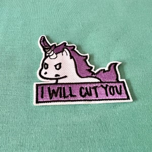 Peut inclure: Patch brodée thermocollante représentant une licorne violette et blanche avec une expression renfrognée et le texte "I will cut you".