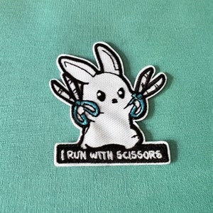 May include: White embroidered patch of a cartoon bunny holding two pairs of blue scissors. The patch has black outlines and the text "I run with scissors" below the bunny.