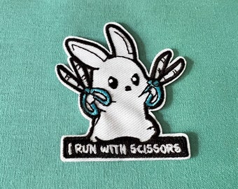 May include: White embroidered patch of a cartoon bunny holding two pairs of blue scissors. The patch has black outlines and the text "I run with scissors" below the bunny.