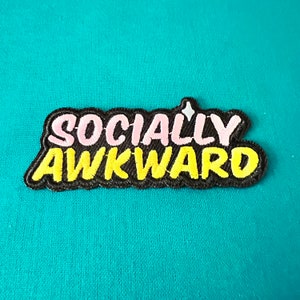 May include: A black embroidered patch with the text "SOCIALLY AWKWARD" in pink and yellow lettering.