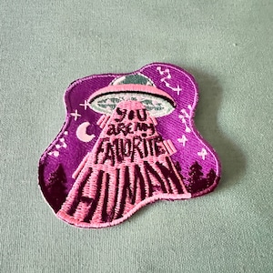 Op de afbeelding: Paarse en roze geborduurde patch met een UFO en de tekst "You are my favorite human".