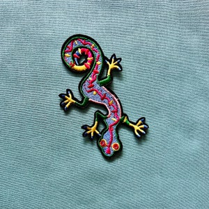Pode incluir: Um remendo bordado colorido de um lagarto verde, azul, rosa e amarelo com a cauda enrolada.