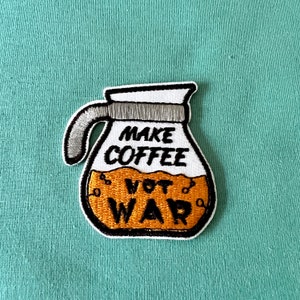 Könnte beinhalten: Weißer gestickter Aufnäher mit einer Kaffeekanne und dem Text "Make Coffee Not War" in Schwarz.