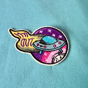 Op de afbeelding: Een kleurrijk geborduurd patch met een UFO die door de ruimte vliegt. De UFO is blauw en grijs met een gele en groene vlam erachter. De tekst "I'm Out" staat op de vlam. De achtergrond is een paarse en roze galaxy met witte sterren.