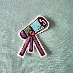 Może przedstawiać: Biała haftowana naszywka przedstawiająca niebieski, zielony i pomarańczowy teleskop na statywie. Teleskop ma czarny kontur, a statyw jest fioletowy.