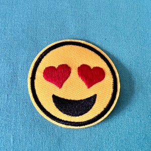 Op de afbeelding: Gele geborduurde smiley met rode hartvormige ogen en een zwarte glimlach.