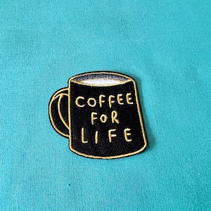 Può includere: Toppa ricamata raffigurante una tazza di caffè nera con la scritta "COFFEE FOR LIFE" in giallo. La tazza ha un manico giallo e un interno bianco, su sfondo turchese. La toppa è destinata a vestiti o accessori.