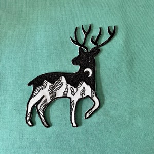 Könnte beinhalten: Schwarz-weiße Silhouette eines Hirsches mit einem Halbmond und einem Gebirge im Inneren seines Körpers. Der Hirsch steht auf einem hellgrünen Hintergrund.