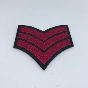 May include: A red and black embroidered patch with three chevrons. The patch is shaped like a chevron and has a black border.