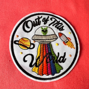 May include: White embroidered patch with a black outline, featuring a cartoon UFO with a rainbow trail, a planet, a rocket, and the text "Out of this World".