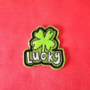 Puede incluir: Un trébol de cuatro hojas verde con un contorno negro y la palabra "LUCKY" en letras blancas con un contorno negro.