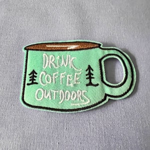 Könnte beinhalten: Grüner gestickter Bügeleisen-Aufnäher in Form einer Kaffeetasse mit dem Text "Drink Coffee Outdoors" und zwei kleinen Kiefern auf beiden Seiten des Textes.