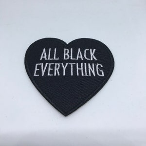 May include: Black heart-shaped patch with white embroidered text reading "ALL BLACK EVERYTHING". The patch is made of a textured fabric and is set against a white background. The patch is a decorative item.