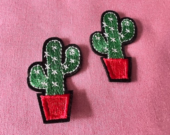 May include: Two green cactus plants with white flowers in red pots. The cactus plants are embroidered on a pink fabric background.