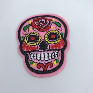 May include: Pink embroidered felt patch of a sugar skull with a red rose, black eyes, and white teeth.