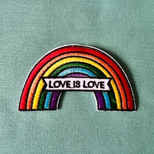 May include: Embroidered rainbow patch with red, orange, yellow, green, blue, and purple stripes. The patch has a white banner with the text "LOVE IS LOVE" in black. The patch is on a light teal fabric.