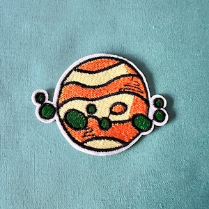 Könnte beinhalten: Ein gestickter Bügel-Patch mit einem Cartoon-Planeten mit orangefarbenen und gelben Streifen und grünen Punkten. Der Planet ist von kleineren grünen Punkten umgeben.