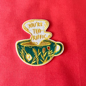 Könnte beinhalten: Eine grüne Teetasse mit einer weißen Dampfwolke darüber. Die Dampfwolke hat den Text "You're Tea-Rific" gestickt. Die Teetasse ist von grünen Blättern und Blumen umgeben.