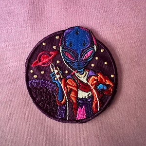 Könnte beinhalten: Ein blauer Außerirdischer mit einem rosafarbenen und roten Planeten im Hintergrund. Der Außerirdische trägt eine rot-orange Jacke und Kopfhörer. Der Außerirdische hält ein Mikrofon in der Hand.