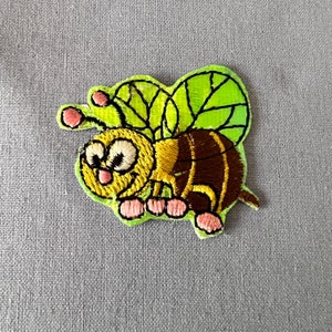 Könnte beinhalten: Eine gestickte Biene in Gelb und Braun mit einem grünen Blattdesign auf dem Rücken. Die Biene hat rosa Füße und ein lächelndes Gesicht.