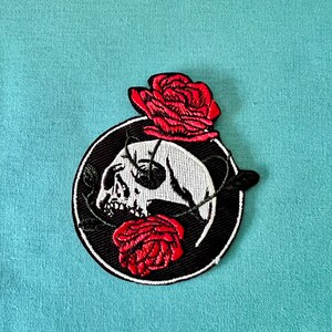 May include: Black and white embroidered skull with red roses on a black background. The skull is surrounded by a vine with leaves and thorns. The roses are in full bloom and have a realistic look.