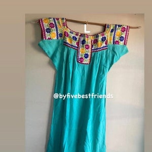 May include: A turquoise blue dress with colorful embroidered flowers around the neckline and sleeves. The dress has short sleeves and a loose fit. The text "@byfivebestfriends" is visible on the dress.