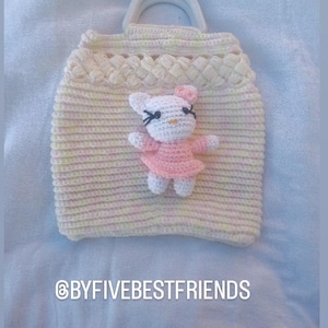 May include: A handmade crochet bag in cream and pastel yellow with a white handle. A small crochet figure of a cat in a pink dress is attached to the front. The text "@BYFIVEBESTFRIENDS" is at the bottom.