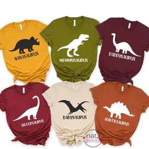 May include: Six t-shirts with different dinosaur silhouettes and text. The shirts are in various colors, including mustard yellow, olive green, burgundy, maroon, beige, and orange. The text on the shirts reads "Nanasaurus", "Mommysaurus", "Daddysaurus", "Sistersaurus", "Babysaurus", and "Auntiesaurus".