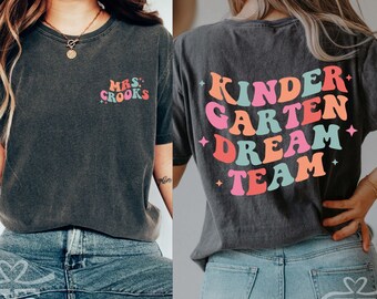 May include: Two women wearing dark gray t-shirts with colorful text. The shirt on the left says "Mrs. Crooks" and the shirt on the right says "Kinder Garden Dream Team". Both women are wearing blue jeans.