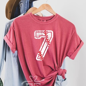 May include: A crimson red t-shirt with a white baseball number 7. The number has a distressed look and is filled with red stitching.