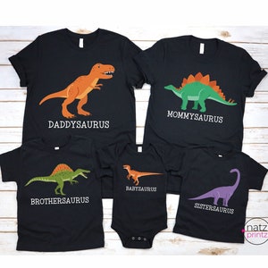 May include: Five black t-shirts with dinosaur graphics and text. The shirts read "Daddysaurus", "Mommysaurus", "Brothersaurus", "Babysaurus", and "Sistersaurus".