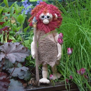 May include: A crocheted plush toy of a caveman with red hair and a beard, holding a wooden spear. The toy is standing in a garden setting with green grass and pink flowers.