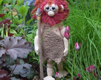 May include: A crocheted plush toy of a caveman with red hair and a beard, holding a wooden spear. The toy is standing in a garden setting with green grass and pink flowers.