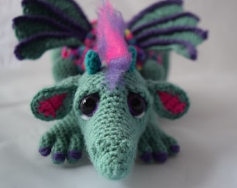 Könnte beinhalten: Ein grüner Häkel-Drache mit lila Flügeln, rosa Ohren und einer rosa-lila flauschigen Mähne. Der Drache hat große schwarze Augen und eine lange Schnauze.