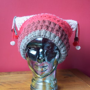 May include: A red, grey, and white crocheted hat with a pointed top and long tassels. The hat is on a mannequin head.