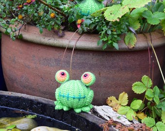 May include: Two green crocheted frogs with orange eyes and stitched mouths. The frogs are sitting on a wooden surface near a pond.