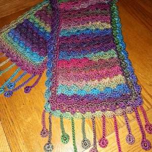 May include: A multi-colored crocheted scarf with a fringe. The scarf is made with a variety of shades of purple, blue, green, and brown yarn.