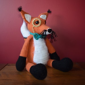 May include: A crocheted fox plush toy with a black monocle, a teal bow tie, and a black mustache. The fox is sitting on a wooden surface.