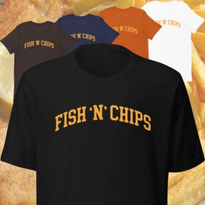 Könnte beinhalten: Schwarzes T-Shirt mit dem Text "FISH 'N' CHIPS" in einem gelben Bogen. Der Text ist in einer fetten, blockartigen Schrift.