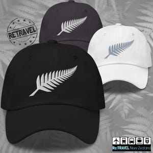 Op de afbeelding: Drie zwarte en witte baseballpetten met een geborduurd zilveren varenbladontwerp. De petten hebben een gebogen klep en een verstelbare band. De tekst "Re:TRAVEL Nieuw-Zeeland" is zichtbaar onderaan de afbeelding.