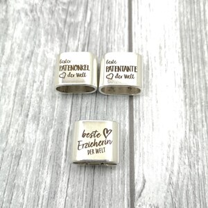 May include: Three silver metal rings with engraved text. The rings say "Beste Patenonkel der Welt", "Beste Patenante der Welt", and "Beste Erzieherin der Welt".