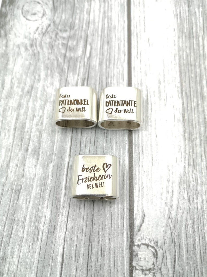 May include: Three silver metal rings with engraved text. The rings say "Beste Patenonkel der Welt", "Beste Patenante der Welt", and "Beste Erzieherin der Welt".