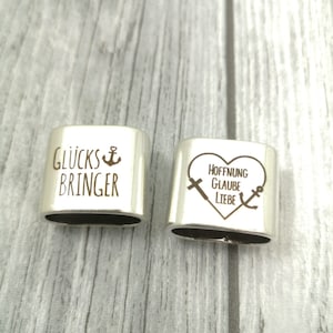 May include: Two silver charm bracelets with engraved messages. The first charm reads "Gl&uuml;cksbringer" with an anchor symbol. The second charm is heart-shaped and reads "Hoffnung Glaube Liebe" with a cross and anchor symbol.