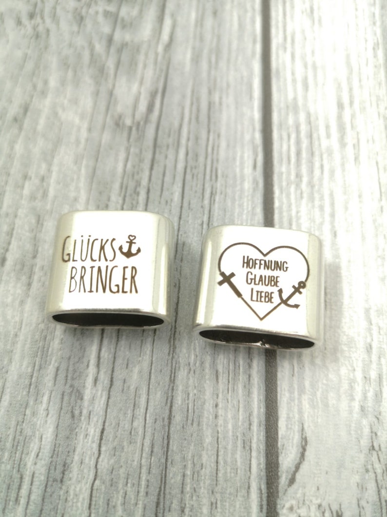 May include: Two silver charm bracelets with engraved messages. The first charm reads "Gl&uuml;cksbringer" with an anchor symbol. The second charm is heart-shaped and reads "Hoffnung Glaube Liebe" with a cross and anchor symbol.