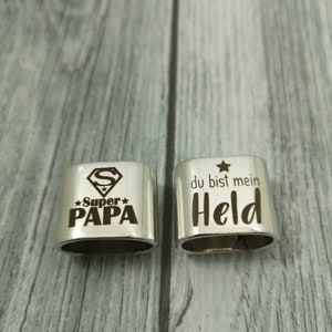 May include: Two silver metal keychains with engraved messages. The first keychain has a Superman logo and the text "Super Papa" with a star on either side. The second keychain has a star and the text "du bist mein Held".