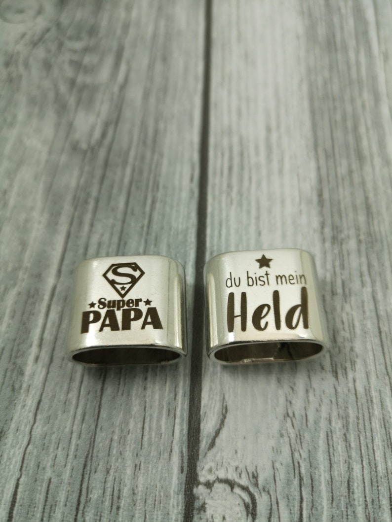 May include: Two silver metal keychains with engraved messages. The first keychain has a Superman logo and the text "Super Papa" with a star on either side. The second keychain has a star and the text "du bist mein Held".