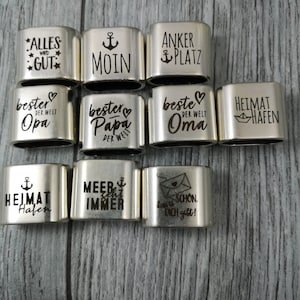 May include: A set of 10 silver metal rings with engraved sayings. The sayings include "Alles wird gut", "Moin", "Anker Platz", "Bester Opa der Welt", "Bester Papa der Welt", "Beste Oma der Welt", "Heimat Hafen", "Heimat Hafen", "Meer sehr immer", and "Das ist schon, dich gibt!"