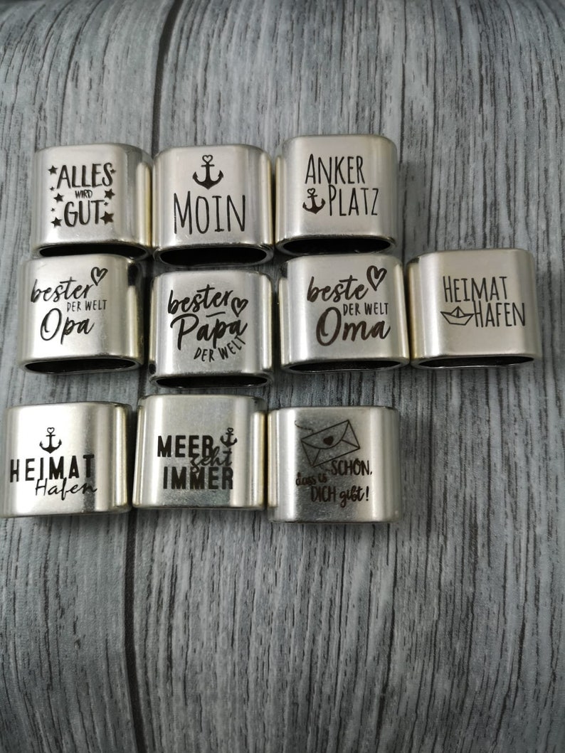 May include: A set of 10 silver metal rings with engraved sayings. The sayings include "Alles wird gut", "Moin", "Anker Platz", "Bester Opa der Welt", "Bester Papa der Welt", "Beste Oma der Welt", "Heimat Hafen", "Heimat Hafen", "Meer sehr immer", and "Das ist schon, dich gibt!"