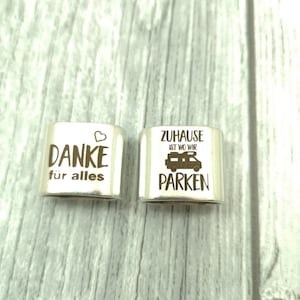 May include: Two silver metal charms with engraved text. The first charm says "Danke f&uuml;r alles" with a heart symbol. The second charm says "Zuhause ist wo wir parken" with a camper van symbol.